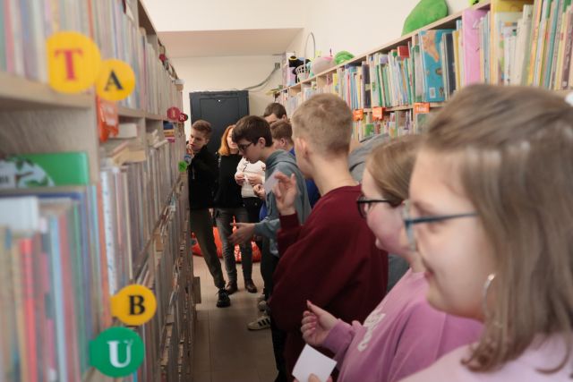Na zdjęciu znajduję się młodzież między regałami z książkami przeglądająca książki na półkach.