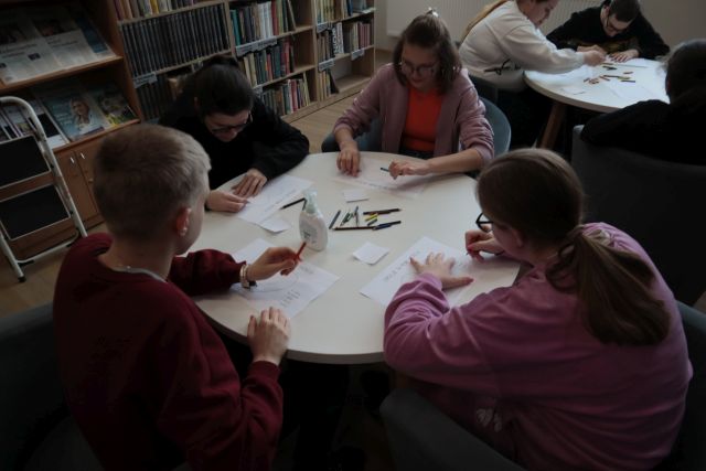 Na zdjęciu znajduję się czwórka młodzieży przy stoliku, która wykonuje prace plastyczną ze znajdujących się na stoliku kartek i kredek. Zajęcia odbywają się w czytelni, po lewej stronie znajdują się półki z książami.