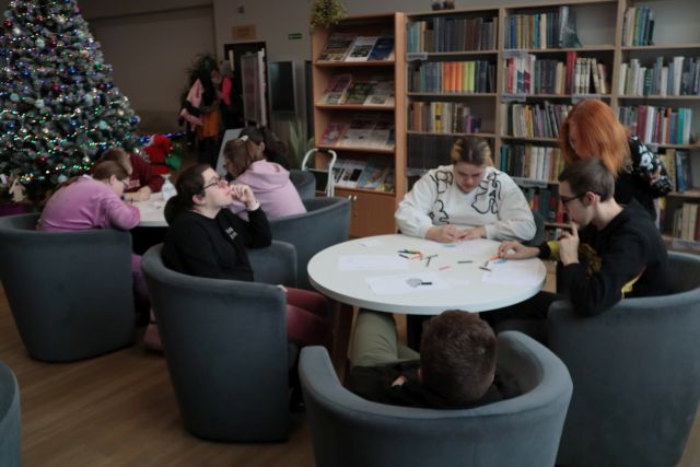 Na zdjęciu znajduję się młodzież przy stolikach, wykonująca prace plastyczną ze znajdujących się na stoliku materiałów. Przy jednym stoiku nachyla się kobieta i przygląda się pracom. W tle znajdują się półki z książkami oraz choinka po lewej stronie.