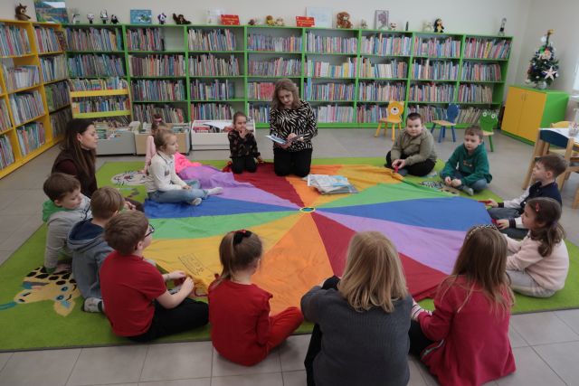Na zdjęciu znajdują się przedszkolaki oraz dwie dorosłe kobiety siedzące dookoła kolorowej chusty. Na środku znajduję się kobieta, która czyta dzieciom książkę. W tle znajdują się półki z książkami.