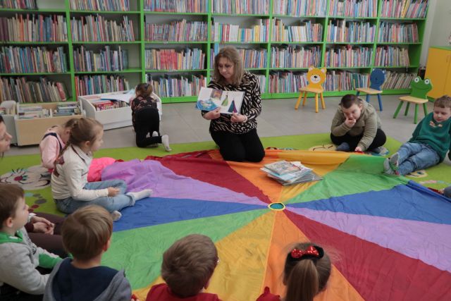 Na zdjęciu znajdują się przedszkolaki siedzące dookoła kolorowej chusty oraz kobieta pokazująca im ilustracje z książki. W tle znajdują się półki z książkami.