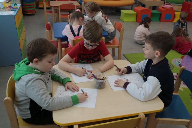 Na zdjęciu znajdują się przedszkolaki przy stolikach kolorująca kolorowanki, na stoliku znajdują się również kredki. W tle znajduję się kolejny stolik, przy którym również siedzą dzieci.
