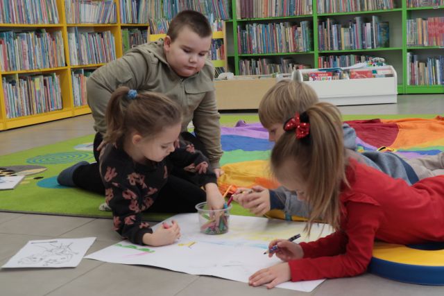 Na zdjęciu znajduję się czwórka dzieci na dywanie kolorujące duży obrazek, jeden chłopiec klęczy i przygląda się pracy. W tle znajdują się półki z książkami.