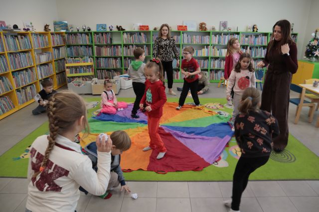 Na zdjęciu znajdują się przedszkola, które rzucają w siebie papierowymi śnieżkami, za nimi oraz po prawej stronie stoją kobiety. W tle znajdują się półki z książkami, a na dywanie leży kolorowa chusta.