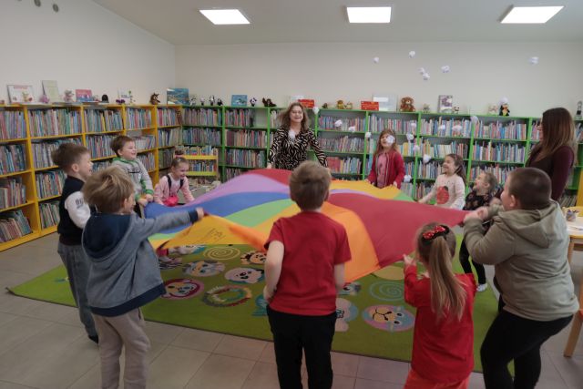 Na zdjęciu znajdują się przedszkolaki oraz kobieta, wszyscy trzymają kolorową chustę, nad którą unoszą się papierowe kulki śniegu. W tle znajdują się półki z książkami.
