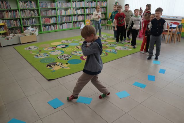 Na zdjęciu znajduję się chłopiec, który idzie z papierowymi kulami śniegu przytulonymi do siebie, omijając niebieskie kartki znajdujące się na podłodze. Po prawej stronie znajdują się dzieci, które czekają na swoją kolej w kolejce. W tle znajduję się dywan oraz półki książkami.