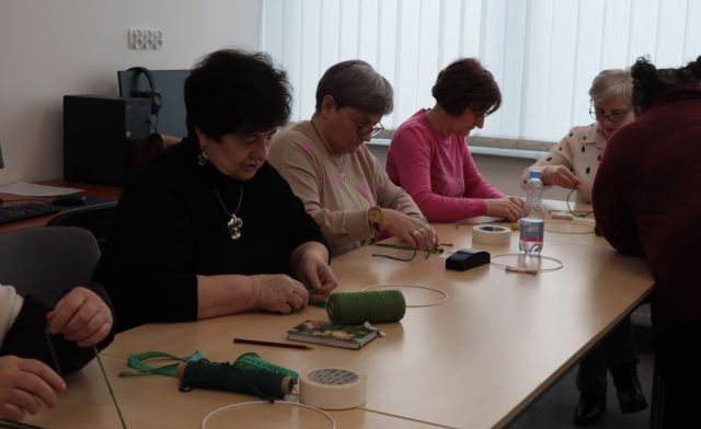 Na zdjęciu znajdują się kobiety siedzące przy stole i wykonujące splot za pomocą makramy. Przed stołem bokiem do kobiet stoi kobieta prowadząca warsztaty. Zajęcia odbywają się w sali komputerowej.
