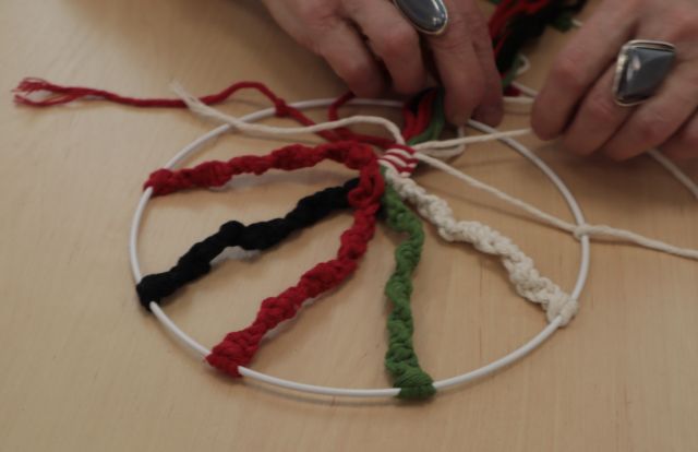 Na zdjęciu znajdują się dłonie kobiety, która robi drzewo życia techniką makramy na metalowym kole.