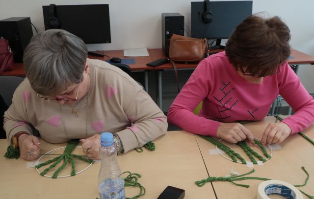 Na zdjęciu znajdują się dwie kobiety wykonujące drzewo życia na kole techniką makramy. Przed kobietą z lewej stoi butelka z wodą, a za kobietami w tle znajdują się komputery.
