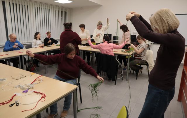 Na zdjęciu znajdują się kobiety stojące przy stołach i ucinające długie paski bawełnianego sznurka. Kobiety znajdują się w sali komputerowej.