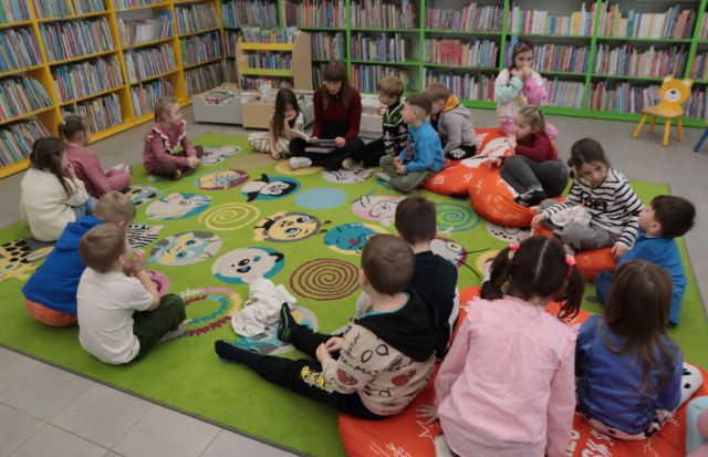 Na zdjęciu znajdują się przedszkolaki siedzące na dywanie, między nimi siedzi kobieta, która czyta im książkę. W tle znajdują się półki z książkami.