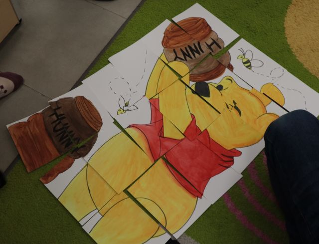 Na zdjęciu znajdują się ułożone, duże puzzle z Kubusia Puchatka, który przechyla nad sobą słoik z miodem, obok znajduję się noga chłopca.