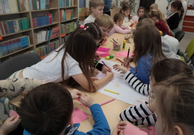 Na zdjęciu znajdują się przedszkolaki przy stole nachylające się nad obrazkiem i kolorujące go. W tle znajdują się półki z książkami.