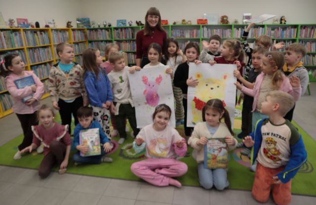 Na zdjęciu znajdują się przedszkolaki, część z nich siedzi na dywanie, część stoi za nimi trzymając dwa duże plakaty, jeden z Kubusiem Puchatkiem, drugi z Prosiaczkiem. Za nimi stoi kobieta. W tle znajdują się półki z książkami.