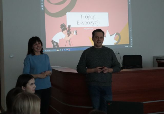 Na zdjęciu znajduję się mężczyzna oraz kobieta po lewej, za nimi znajduję się biurko biurowe, a na ścianie wyświetlana jest prezentacja o Trójkącie Ekspozycji. Przed kobietą od boku znajdują się trzy głowy.