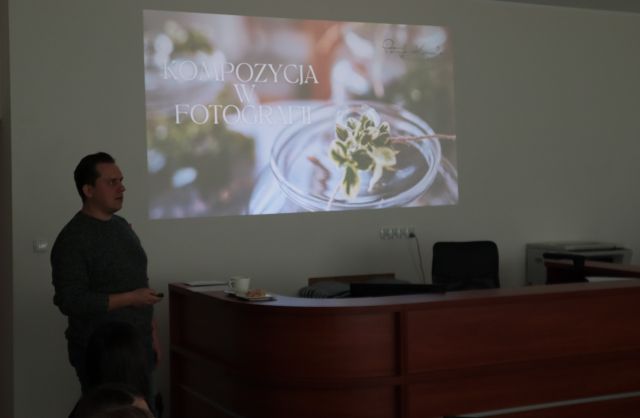 Na zdjęciu po lewej stronie znajduje się mężczyzna stojący przy biurku. Na ścianie za nim wyświetlana jest prezentacja dotycząca Kompozycji w fotografii.