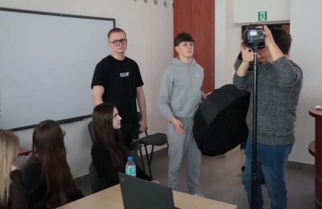 Na zdjęciu znajduję się piątka młodzieży, trzy dziewczyny siedzą przy stole, na którym leży laptop. Koło dziewczyn stoi dwójka chłopaków, jeden z nich trzyma czarny materiał w ręce i są skierowni w stronę mężczyzny po prawej, który trzyma aparat znajdujący się na statywie. Po lewej stronie znajduję się tablica interaktywna.