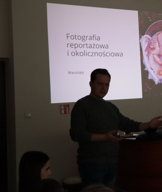 Na zdjęciu znajduję się mężczyzna od pasa w górę, który opiera się o biurko, a za nim wyświetlana jest prezentacja o Fotografii reportażowej i okolicznościowej.
