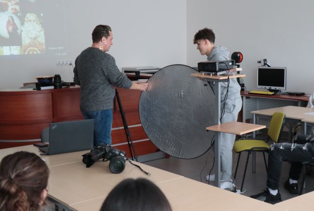 Na zdjęciu znajduje się chłopak trzymający blendę fotograficzną, obok niego stoi mężczyzna i trzyma ją jedną ręką. Przed nimi stoi projektor, a dookoła stoją stoły, przy których siedzi młodzież.. Warsztaty odbywają się w sali komputerowej.