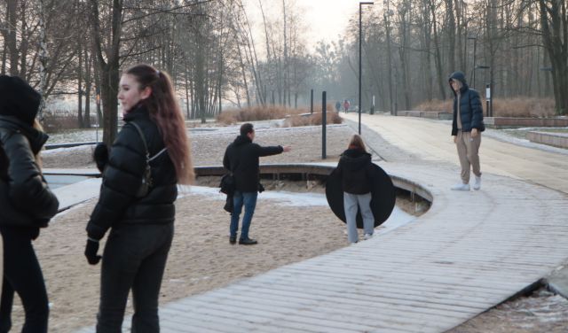Na zdjęciu po lewej stronie znajdują się dwie ubrane w kurtki dziewczyny, w tle znajduje sie chłopak pozujący do zdjęcia na kawałku okrągłego pomostu, przed nim stoi dziewczyna z blendą fotograficzną oraz fotograf z wyciągniętą ręką. Młodzież znajduje się nad jeziorem przy lesie.