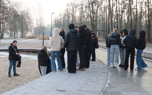 Na zdjęciu znajduję się grupka młodzieży, która stoi na podwyższeniu, na dole na pisaku stoi mężczyzna z aparatem oraz dziewczyna z blendą fotograficzną. Warsztaty fotograficzne odbywają się nad jeziorem przy lesie.