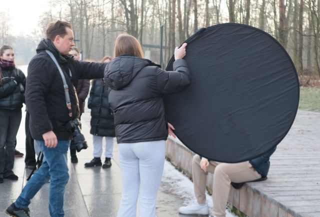 Na zdjęciu znajduję się dziewczyna trzymająca blendę fotograficzną, za którą siedzi chłopak, obok dziewczyny stoi fotograf. W tle znajduje się młodzież obserwująca warsztaty, które odbywają się nad jeziorem przy lesie.