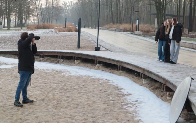 Na zdjęciu znajduje się fotograf stojący na piasku przed pomostem, na którym stoi trójka młodzieży. Przed pomostem znajduje się trochę śniegu, a w tle znajduje się las.