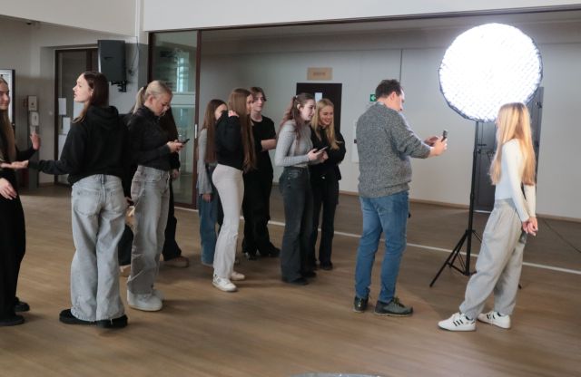 Na zdjęciu znajduje sie grupka młodzieży przyglądająca się dziewczynie, której fotograf robi zdjęcie telefonem. Obok dziewczyny stoi zapalona lampa fotograficzna.