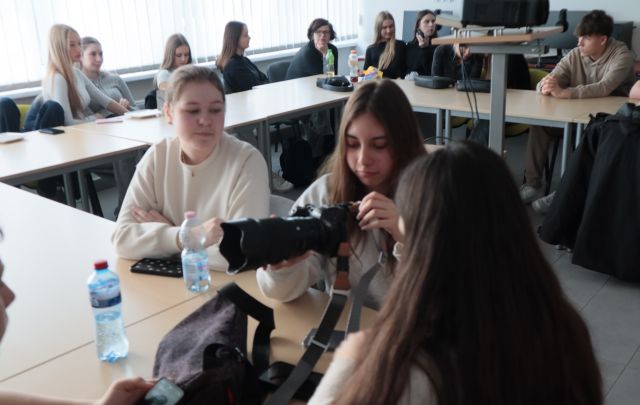 Na zdjęciu znajduje się młodzież przy złączonych stołach w literę C, w centrum znajduje się dziewczyna oglądająca trzymany w dłoniach aparat. Warsztaty odbywają się w sali komputerowej.