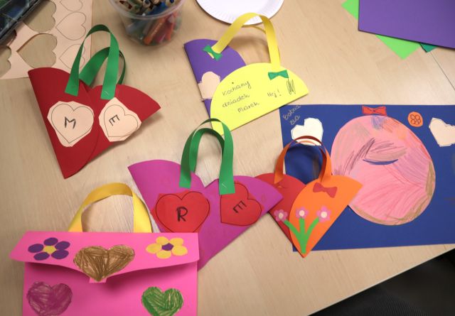 Na zdjęciu znajdują sie kolorowe, papierowe prace, cztery koszyki, kopertka i laurka. Wszystkie prace są ozdobione sa kolorowymi serduszkami, kwiatkami. Prace znajdują się na stole i dookoła znajdują się kolorowe wycinki.