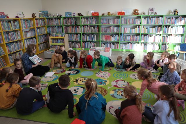 Na zdjęciu znajdują się przedszkolaki siedzące w okręgu na dywanie oraz kobieta, która pokazuje im ilustracje książki. W tle znajdują się półki z książkami.