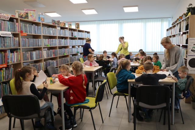 Na zdjęciu znajdują się przedszkolaki siedzące przy stolikach , przy których wykonują prace plastyczną z białych kartek, między nimi stoją trzy kobiety. W tle znajdują się okna, a po bokach półki z książkami.