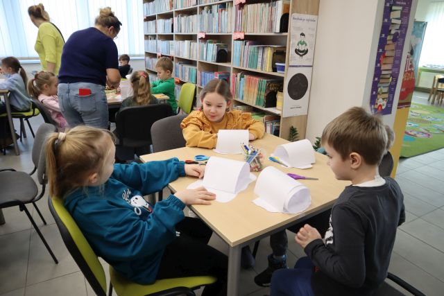 Na zdjęciu znajduje się czwórka dzieci przy stoliku, na którym wykonują igloo z białych kartek oraz znajdujących się na stole kredek. W tle znajduje się reszta przedszkolaków przy stolikach oraz nachylające się przy nich Panie. W tle znajdują się okna oraz półki z książkami.