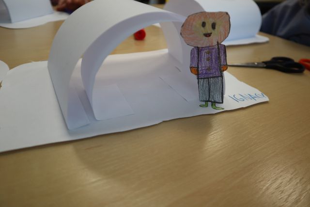Na zdjęciu znajduje się papierowe igloo wraz z kolorowym rysunkiem chłopca wykonane przez przedszkolaka. W tle na stoliku znajdują się dwie inne prace.