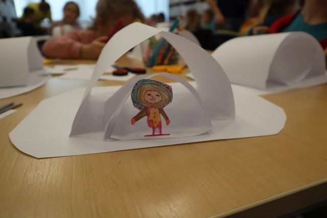 Na zdjęciu znajduje sie białe papierowe igloo wykonane przez przedszkolaka, przed nim stoi mały papierowy mieszkaniec igloo, w tle znajdują się inne prace.