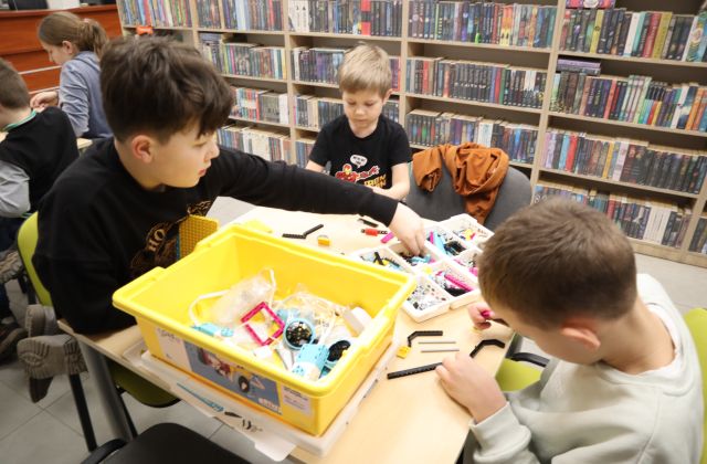 Na zdjęciu znajduje się trójka chłopców przy stoliku, jeden z nich nachyla się aby sięgnąć coś z końca. Na stoliku znajduje się zestaw LEGO Education, z którego chłopcy budują rakietę. W tle znajdują się półki z książkami.