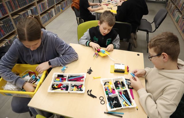 Na zdjęciu przy stoliku znajduję się dwóch chłopców oraz jedną dziewczynę. Chłopcy budują rakietę z LEGO Education, które znajduje się na stole, a dziewczyna trzyma na kolach pudełko, w którym szuka elementów. W tle znajduje się jeszcze jeden stolik, a dookoła półki z książkami.