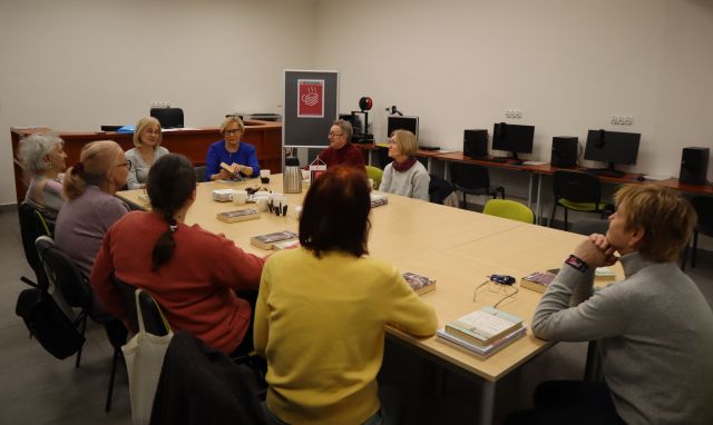 przy połączonych stołach siedzi 9 kobiet, przed nimi na stole proporczyk DKK, kubki i termosy, w tle biurka z komputerami