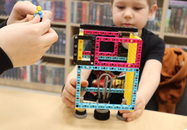 Na zdjęciu znajduje się chłopiec trzymający zbudowaną rakietę z LEGO Education, po lewej stronie znajdują się dziecięce ręce dokładające element do budowli. W tle znajdują się półki z książkami.