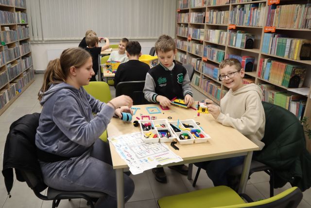 Na zdjęciu znajdują się dwa stoliku, jeden za drugim, przy których siedzą po trzy osoby i budują rakietę z LEGO Education znajdującego się na stole. W tle znajdują się okna, a po bokach półki z książkami.