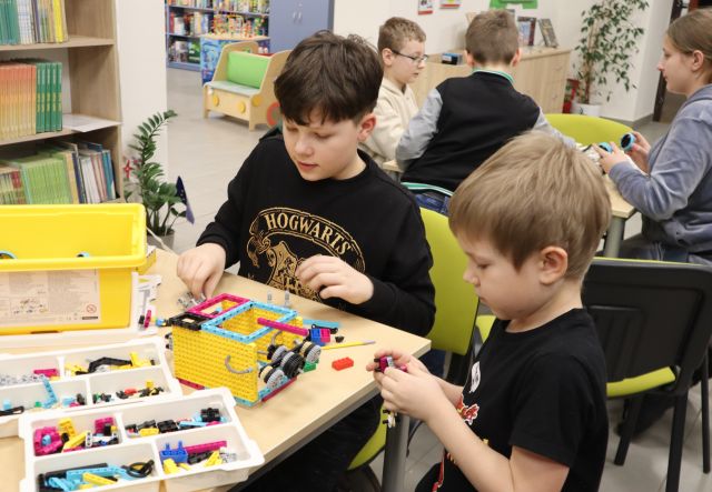 Na zdjęciu znajduje się dwójka chłopców przy stoliku, na którym leży zestaw LEGO Education, za nimi znajduję się trójka osób przy osobnym stoliku. W tle znajdują się półki z książkami.