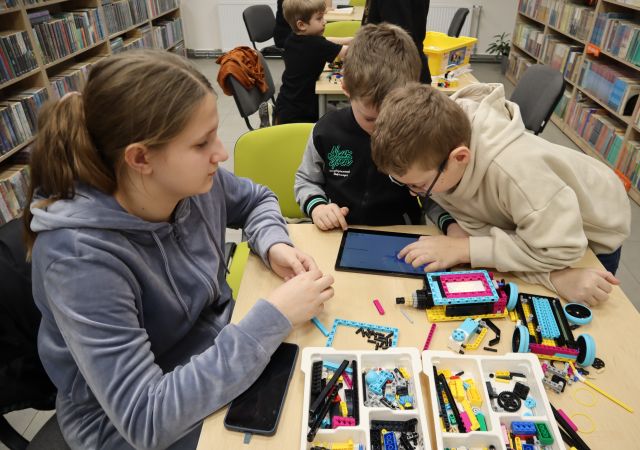 Na zdjęciu znajduje się trójka osób przy stoliku, jeden chłopiec nachyla się nad chłopcem obok, który trzyma przed sobą tablet. Na stoliku znajdują się klocki LEGO Education. W tle znajduje się dwójka chłopców przy osobnym stoliku, a po bokach półki z książkami.