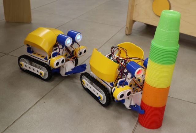 Na zdjęciu znajdują się dwa roboty Skribot, jeden jedzie za drugim. Pierwszy z nich trzyma kolorowe plastikowe kubki.
