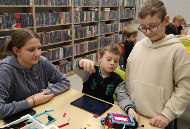 Na zdjęciu znajduje się dziewczynka oraz dwóch chłopców, przed jednym leży tablet i wyciąga on rękę w bok. W tle znajdują się półki z książkami.