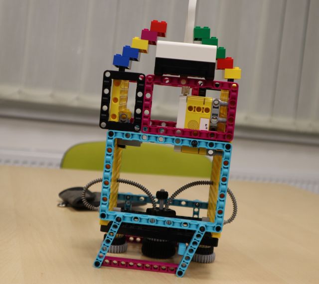 Na zdjęciu znajduje się kwadratowy prototyp rakiety zbudowanej z LEGO, w tle znajduje się kawałek krzesła oraz okna.