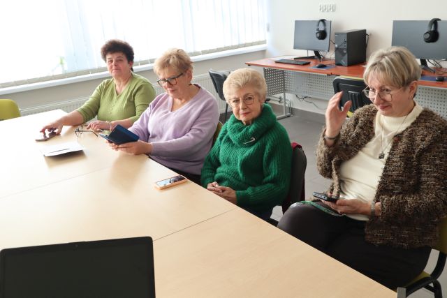 4 seniorki siedzą przy stołach, w rękach mają telefony komórkowe, w tle biurka z komputerami