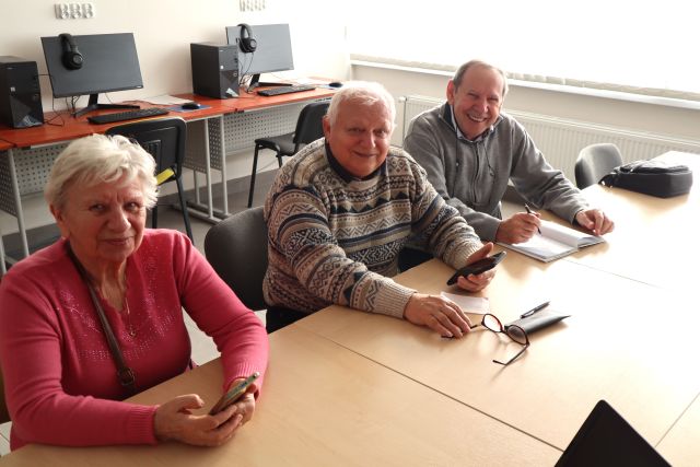 3 seniorów siedzi przy stołach z telefonami komórkowymi, uśmiechają się do aparatu. w tle biurka z komputerami