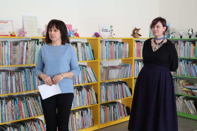 Na zdjęciu znajduje się kobieta w czarnej sukience, po lewej stoi kobieta w niebieskiej bluzce i trzyma w rękach przed sobą kartkę. W tle znajdują się półki z książkami.