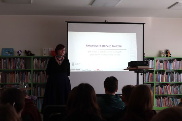 Na zdjęciu znajduje się kobieta stojąca obok ekranu, na którym wyświetlana jest prezentacja za pomocą stojącego przed nim rzutnika. Przed kobietą siedzi młodzież. W tle znajdują się półki z książkami.