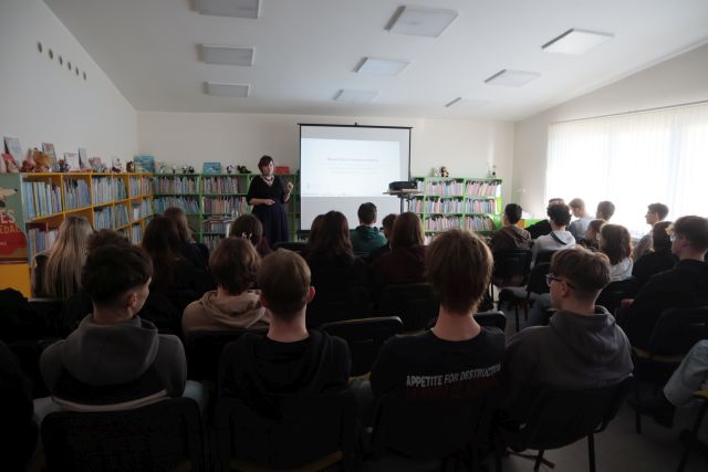 Na zdjęciu znajduje się młodzież na krzesłach skierowana w stronę kobiety, która przedstawia prezentację na temat Ekosłowiańskości. Po lewej stronie znajdują się półki z książkami, a po prawej okno.